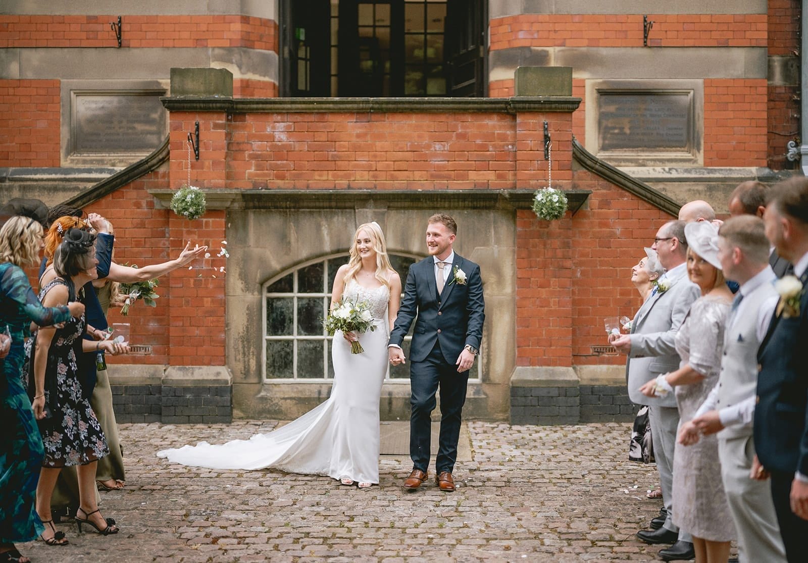 A bride and groom stand smiling on a cobblestone pathway in front of a brick building. Guests on either side throw confetti and clap. The bride wears a white dress and holds a bouquet, while the groom is in a dark suit. Everyone appears joyful.
