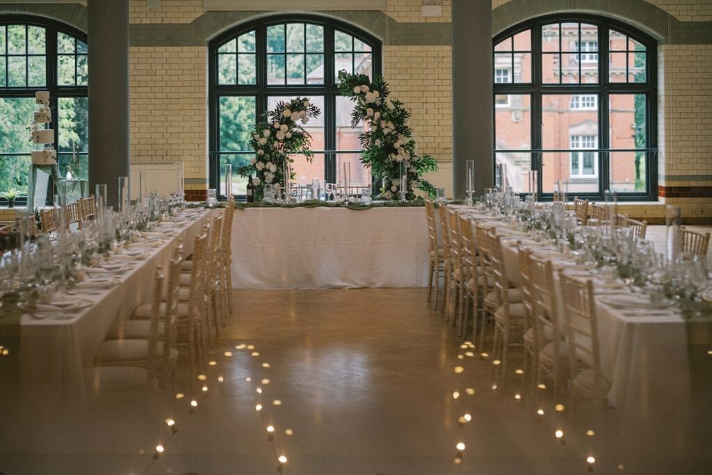 A beautifully arranged wedding reception hall with two long tables filled with white tableware and chairs. In the background, a floral arch sits in front of large arched windows, providing a view of greenery and the red brick charm perfect for a Pumping House wedding photographers lens.