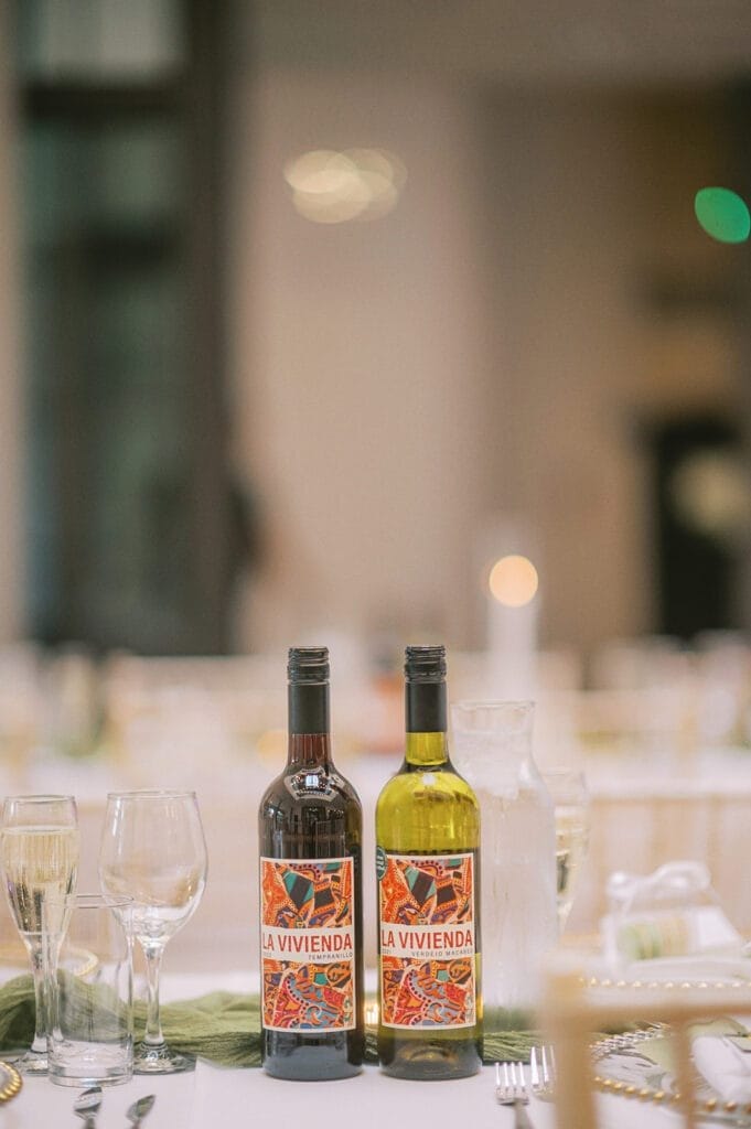 Two wine bottles labeled La Vivienda, one red and one white, grace a table set for dining, perhaps captured by a keen wedding photographer. The setting features glasses, cutlery, and a green cloth runner, with a softly blurred backdrop suggesting a cozy restaurant ambiance.
