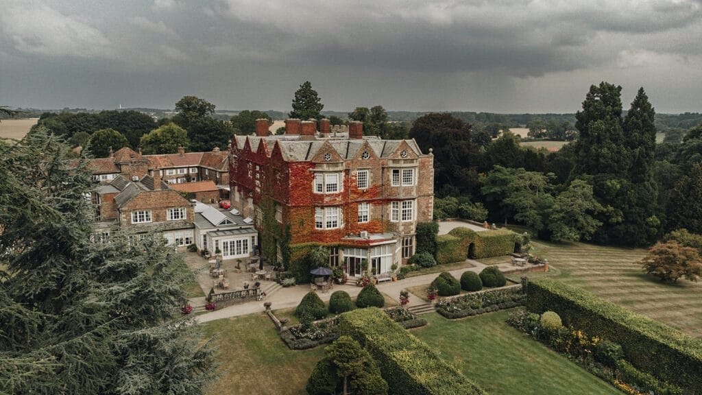Goldsborough Hall Wedding | Yorkshire Wedding Photographer 2 An aerial view captures the majesty of a large, historic stone mansion, its ivy-covered walls whispering tales of old. Surrounded by well-manicured gardens and lush trees under a cloudy sky, this scene is a dream canvas for any Goldsborough Hall wedding photographer seeking countryside charm.