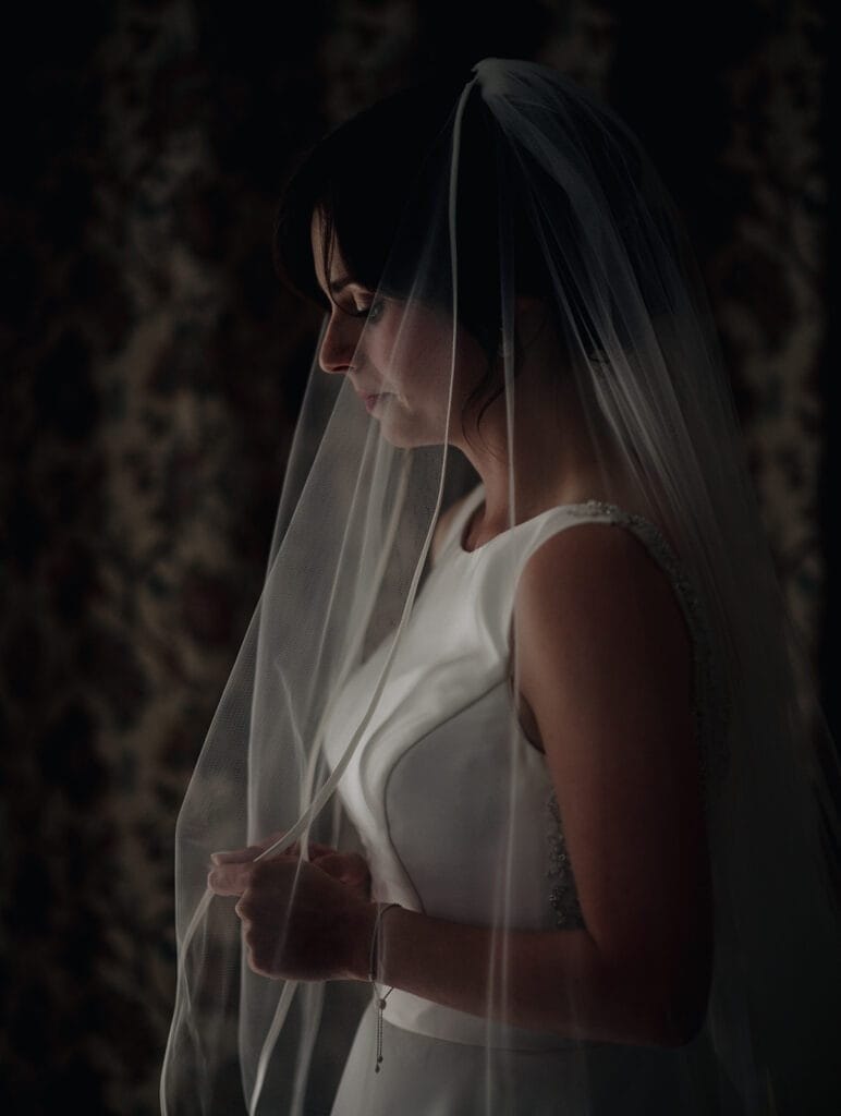 Goldsborough Hall Wedding | Yorkshire Wedding Photographer 17 A bride in a white dress stands in profile, holding her veil. The dim lighting creates a soft, intimate atmosphere as she gazes downward. Captured by a Goldsborough Hall wedding photographer, the moment exudes timeless elegance and grace.