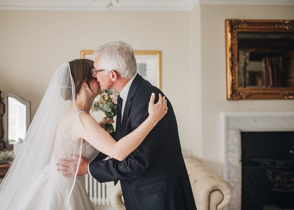 Goldsborough Hall Wedding | Yorkshire Wedding Photographer 21 A bride in a white dress and veil embraces an older man, likely her father, in a warmly lit room at Goldsborough Hall. The scene is beautifully framed with artwork and a fireplace, captured perfectly by the wedding photographer.
