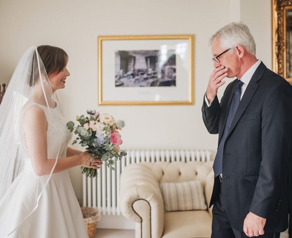 Goldsborough Hall Wedding | Yorkshire Wedding Photographer 22 A bride in a white gown and veil holds a bouquet, smiling at an older man in a suit whos touching his face emotionally. Captured by a seasoned Goldsborough Hall wedding photographer, they stand in a room with a beige couch and framed artwork on the wall.