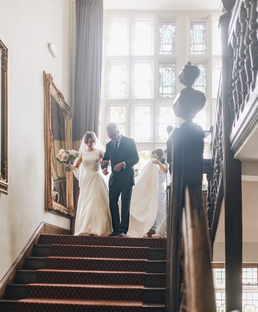 Goldsborough Hall Wedding | Yorkshire Wedding Photographer 23 A bride in a white gown gracefully descends the staircase at Goldsborough Hall, an older man by her side. She clutches a bouquet as attendants adjust her veil. With large windows and framed paintings lining the carpeted stairs, this moment is beautifully captured by the wedding photographer.