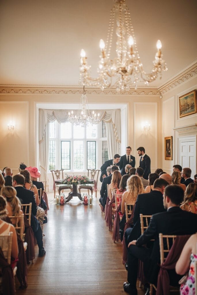 Goldsborough Hall Wedding | Yorkshire Wedding Photographer 25 A wedding ceremony in a bright, elegant room with chandeliers and large windows captures the eye, perfect for a Goldsborough Hall wedding photographer. Guests sit facing the groom and groomsmen near a floral-adorned table. The venues ornate decorations and soft lighting enhance the ambiance.