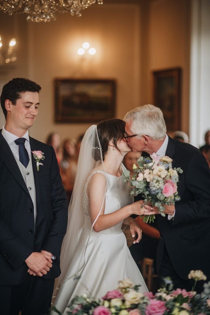 Goldsborough Hall Wedding | Yorkshire Wedding Photographer 29 In a beautifully adorned room at Goldsborough Hall, a bride in a white dress and veil kisses an older man while holding her bouquet. Beside them, a younger man in a suit smiles warmly as guests look on, capturing the magic through the lens of their wedding photographer.