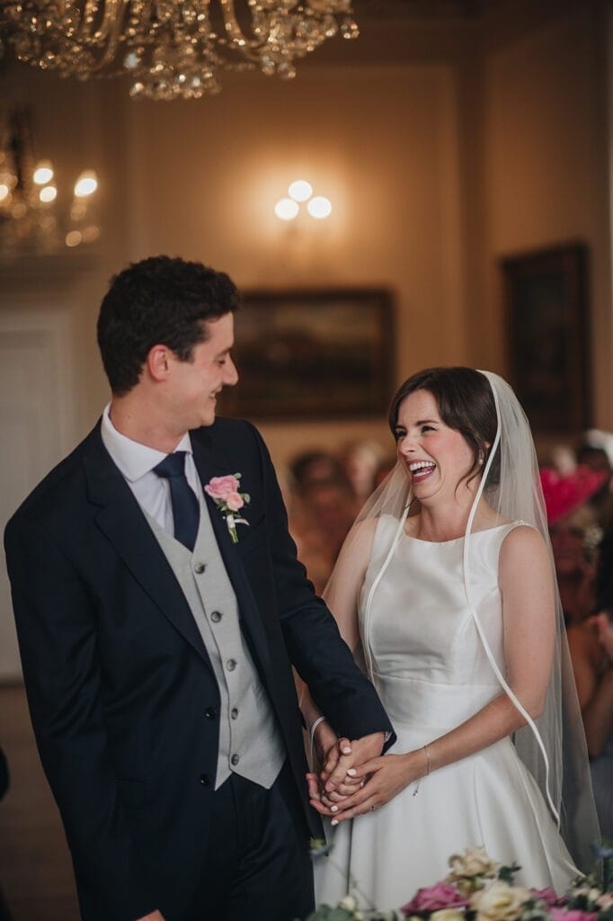 Goldsborough Hall Wedding | Yorkshire Wedding Photographer 39 A bride and groom smile joyfully at each other during a Goldsborough Hall wedding ceremony. The bride is in a white dress and veil, while the groom sports a dark suit with a pink boutonniere. Holding hands beneath the chandeliers, their moment is perfectly captured by the wedding photographer.