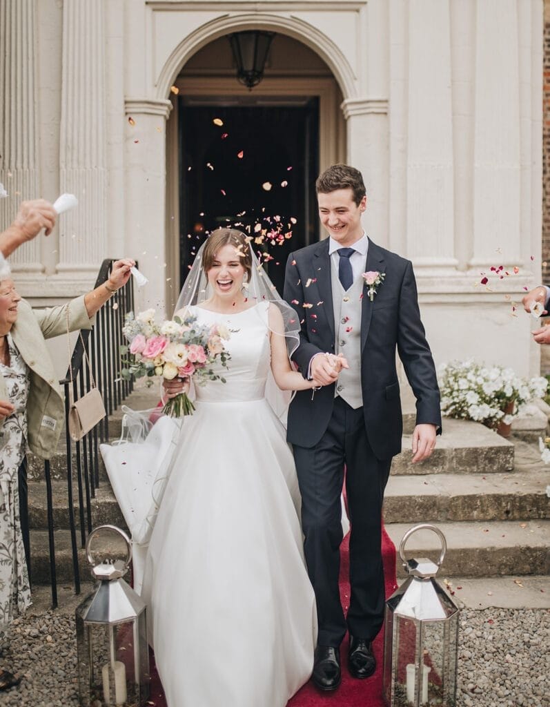 Goldsborough Hall Wedding | Yorkshire Wedding Photographer 43 The joyful bride and groom walk hand in hand down a red carpet at Goldsborough Hall, enveloped by the lens of a wedding photographer. The bride clutches her bouquet as guests shower them with flower petals. Large lanterns beautifully adorn the steps, adding to the enchanting scene.