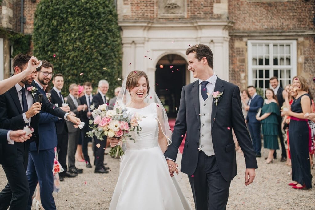 Goldsborough Hall Wedding | Yorkshire Wedding Photographer 44 A joyful bride and groom exit a historic brick building, hand in hand, while guests cheer and throw confetti. The bride holds a bouquet of pink and white flowers, both smiling brightly. Capturing this moment is your skilled Goldsborough Hall wedding photographer amid guests in formal attire.