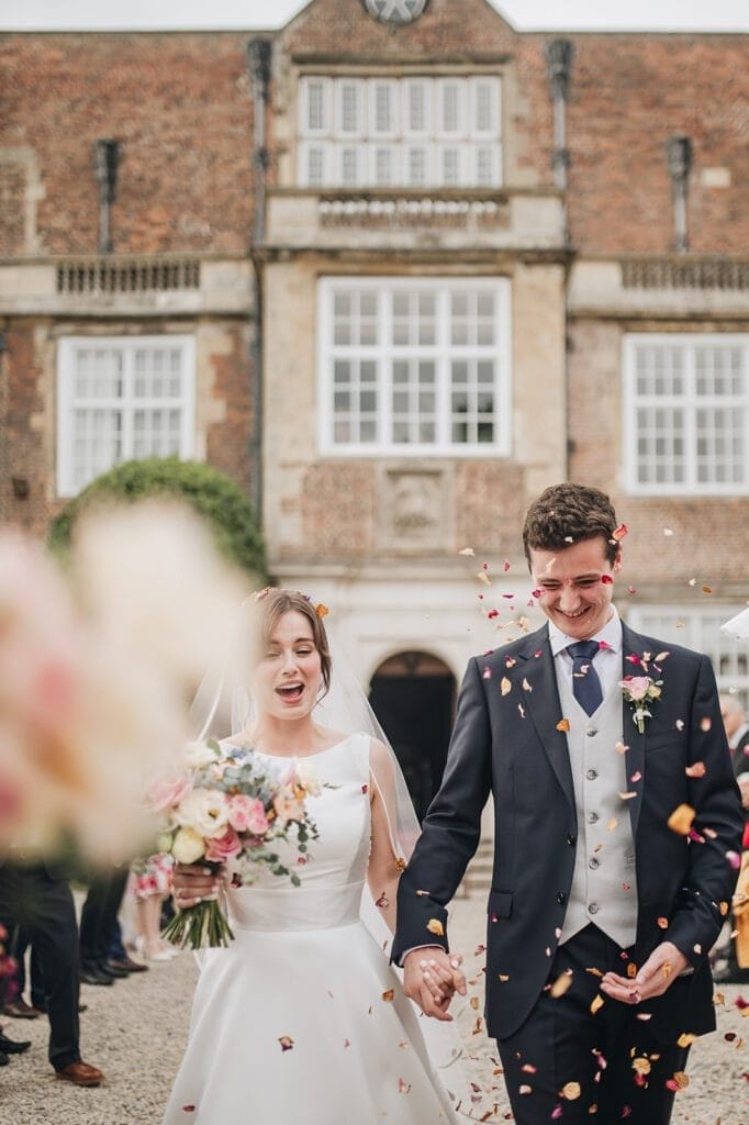 Goldsborough Hall Wedding | Yorkshire Wedding Photographer 47 A joyful couple in wedding attire walk hand in hand outside a brick building. The bride, holding a bouquet, wears a white dress and veil, while the groom is in a dark suit. Captured by an expert Goldsborough Hall wedding photographer, they smile as petals are showered over them.