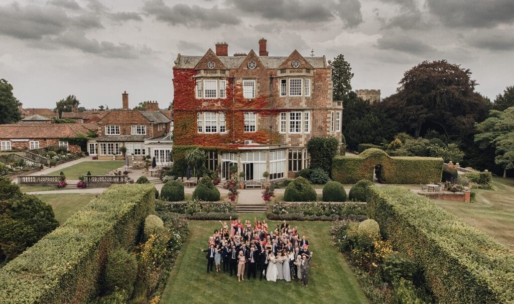 Goldsborough Hall Wedding | Yorkshire Wedding Photographer 51 A group of people gathered for a wedding celebration in front of an ivy-covered historic mansion. Captured by a Goldsborough Hall wedding photographer, the scene is surrounded by manicured gardens and hedges, under a cloudy sky.