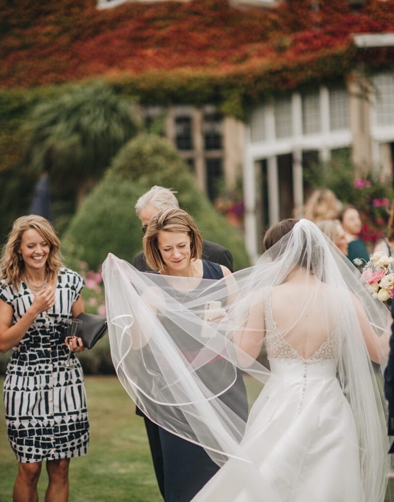 Goldsborough Hall Wedding | Yorkshire Wedding Photographer 52 At a Goldsborough Hall wedding, the bride in a white dress and veil mingles with guests at an outdoor event. Two women smile—one in a patterned dress, the other holding a drink. Lush greenery and flowers surround the picturesque building in the background.