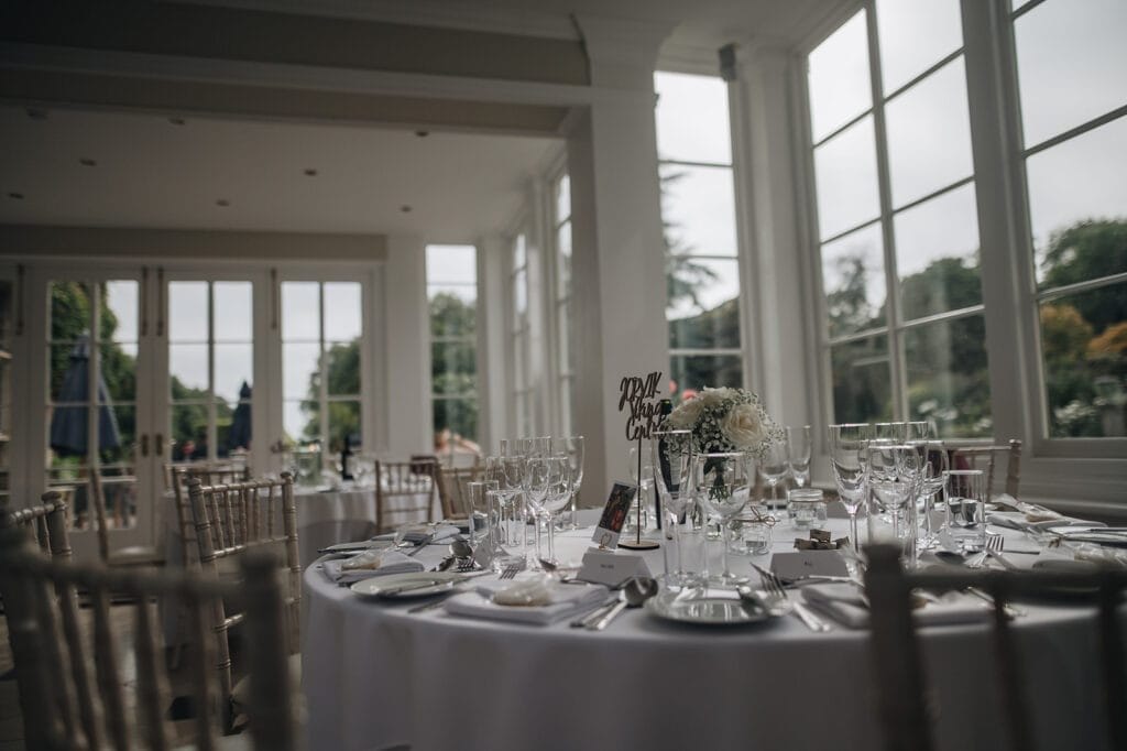 Goldsborough Hall Wedding | Yorkshire Wedding Photographer 59 A round wedding reception table is elegantly set with a white tablecloth, glassware, place settings, and a floral centerpiece. In the bright room with large windows revealing a garden view, every moment deserves to be captured by a Goldsborough Hall wedding photographer.