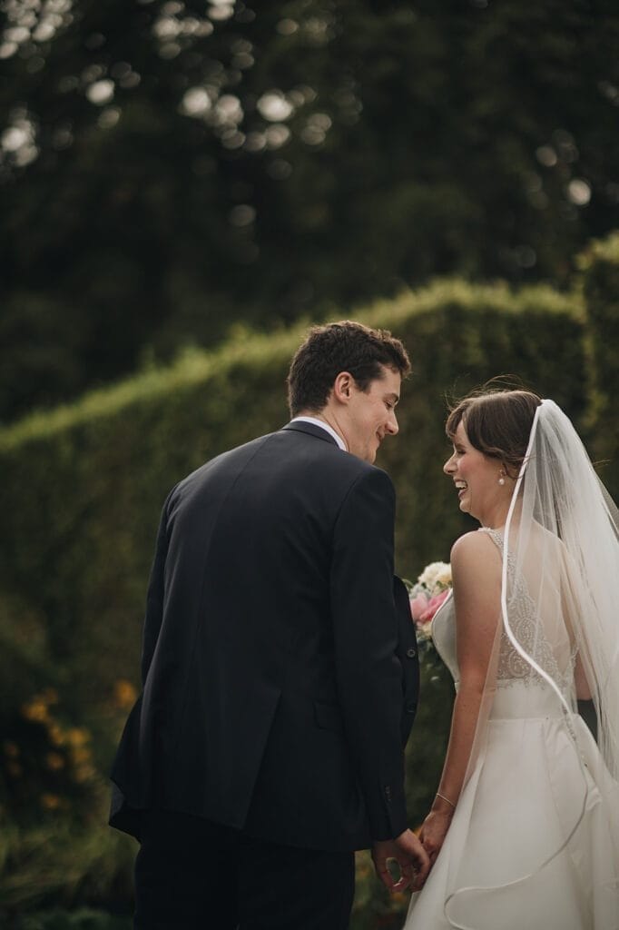 Goldsborough Hall Wedding | Yorkshire Wedding Photographer 65 A bride and groom hold hands and smile at each other in a lush garden setting at Goldsborough Hall. The bride wears a white dress with a veil, the groom in a dark suit. Their wedding photographer captures the moment, as the greenery adds a natural, romantic atmosphere.