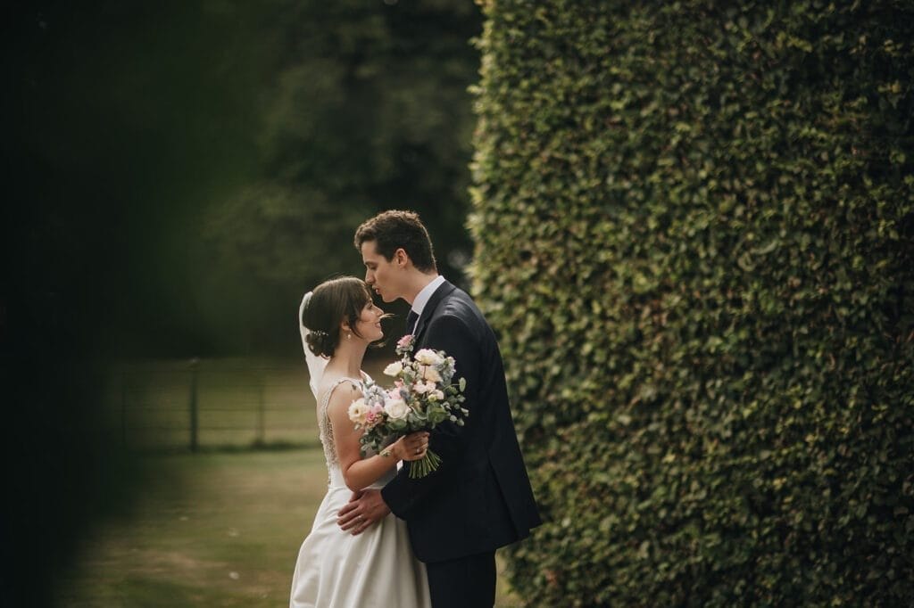 Goldsborough Hall Wedding | Yorkshire Wedding Photographer 64 A bride and groom stand closely beside a tall hedge at Goldsborough Hall. The groom kisses the brides forehead as she holds a bouquet of flowers. They are both in wedding attire, captured beautifully by their wedding photographer against the lush garden setting.