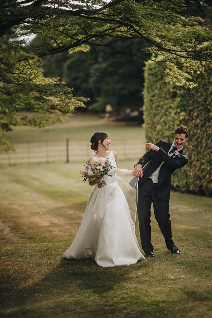 Goldsborough Hall Wedding | Yorkshire Wedding Photographer 72 At Goldsborough Hall, a bride and groom joyously pose under a grand tree in the grassy garden, surrounded by neatly trimmed hedges. The brides laughter fills the air as she holds her bouquet while the groom playfully points away, both appearing happy and relaxed.