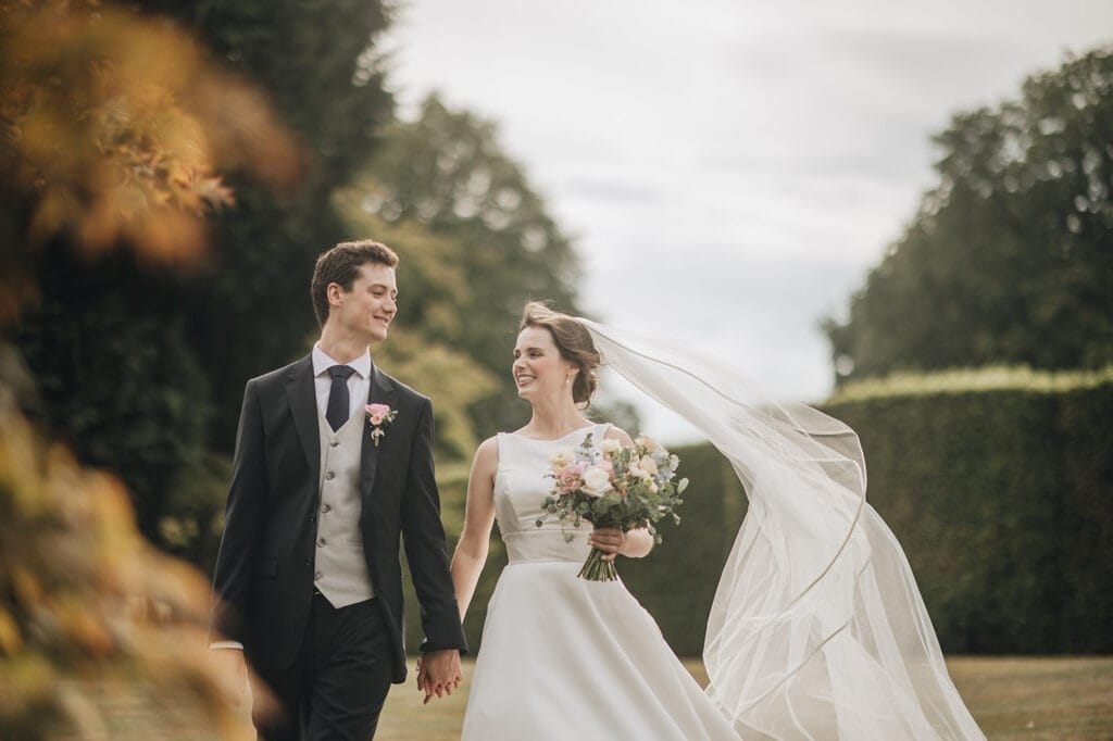 Goldsborough Hall Wedding | Yorkshire Wedding Photographer 69 A bride and groom, smiling and holding hands, walk outside at Goldsborough Hall. The brides white dress with a long veil flows in the wind as she holds a bouquet. The groom is in a dark suit and vest. Greenery and blurred trees create a serene backdrop for this magical moment captured by their wedding photographer.