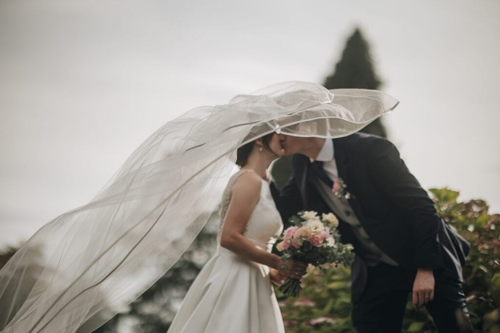 Goldsborough Hall Wedding | Yorkshire Wedding Photographer 73 A couple kisses outdoors on their wedding day at Goldsborough Hall. The brides flowing veil catches the wind as she holds a bouquet of flowers, and the groom stands beside her in a dark suit. Greenery fills the blurred background, captured beautifully by their photographer.