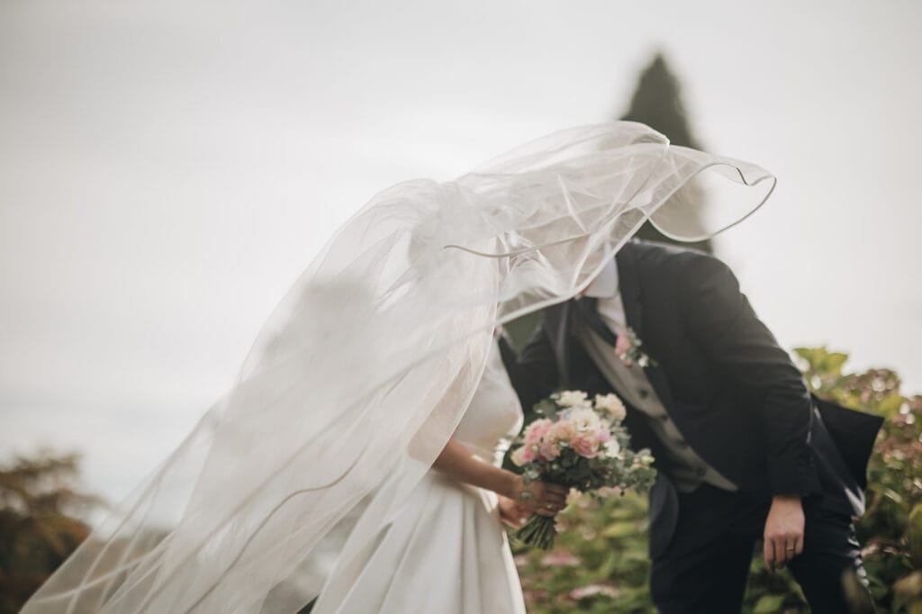 Goldsborough Hall Wedding | Yorkshire Wedding Photographer 76 A bride and groom share a tender kiss outdoors at Goldsborough Hall, her veil billowing dramatically in the breeze. She clasps a bouquet of pink and white flowers, set against lush greenery and a tree under a cloudy sky captured by the wedding photographer.