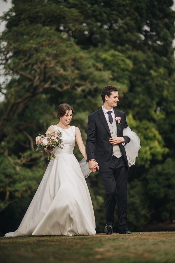 Goldsborough Hall Wedding | Yorkshire Wedding Photographer 71 A bride in a white gown and a groom in a black suit walk hand in hand across the grass, beaming with joy. The bride clutches a bouquet and drapes a veil, embodying elegance against the lush green trees—a scene perfectly captured by Goldsborough Hall wedding photographer.JPG.