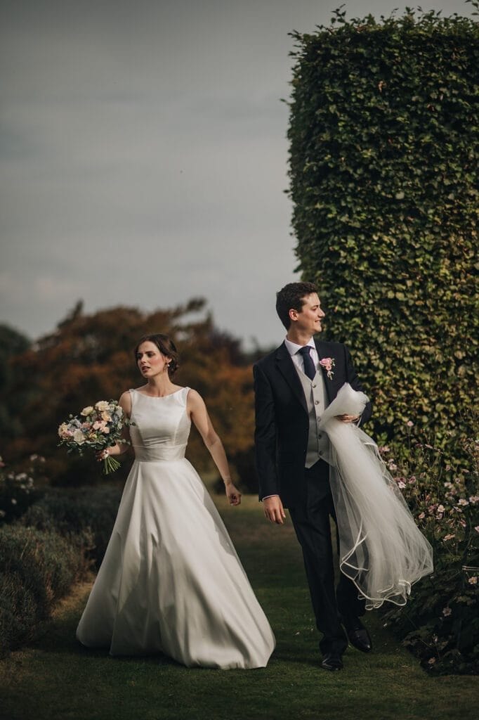 Goldsborough Hall Wedding | Yorkshire Wedding Photographer 63 A bride and groom walk through the garden path at Goldsborough Hall. The bride, in a white gown and holding a bouquet, is accompanied by her groom in a dark suit, who carries her dress train. Theyre surrounded by vibrant greenery and flowers, with a tall hedge completing this enchanting moment.