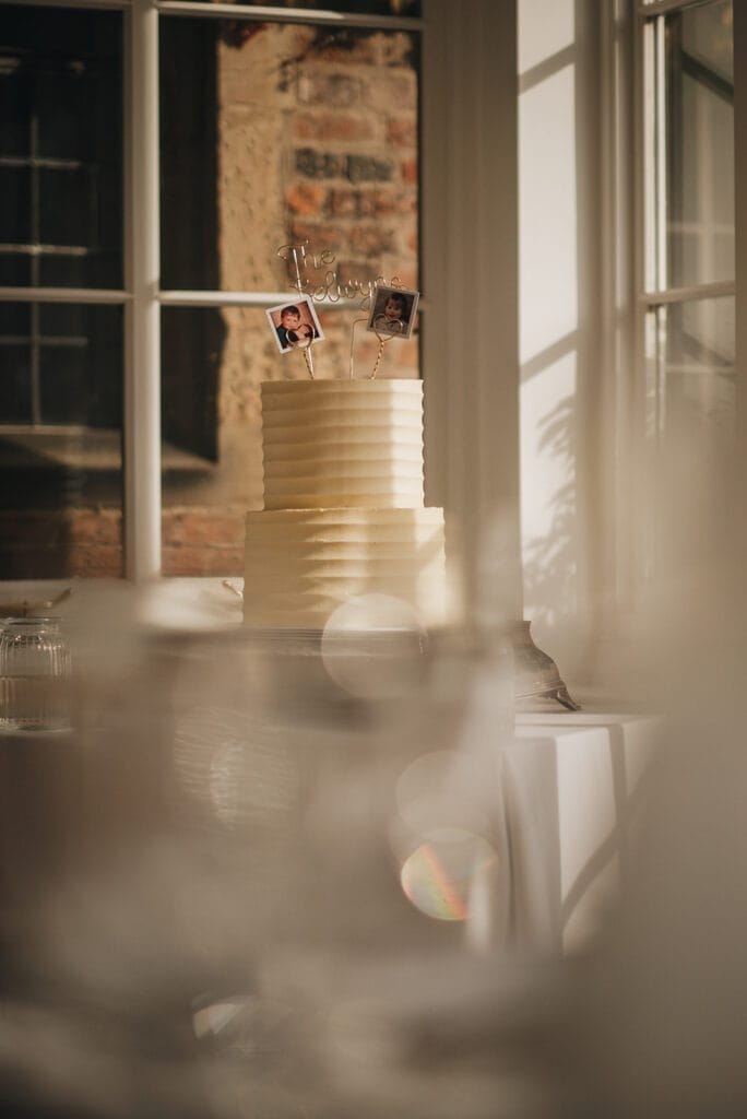 Goldsborough Hall Wedding | Yorkshire Wedding Photographer 62 A tiered white cake with ribbed texture is elegantly showcased in a sunlit room, perfect for any Goldsborough Hall wedding photographer to capture. It features photo toppers and a script word decoration, while a window reveals a glimpse of the brick wall outside. Blurred elements frame this exquisite scene.