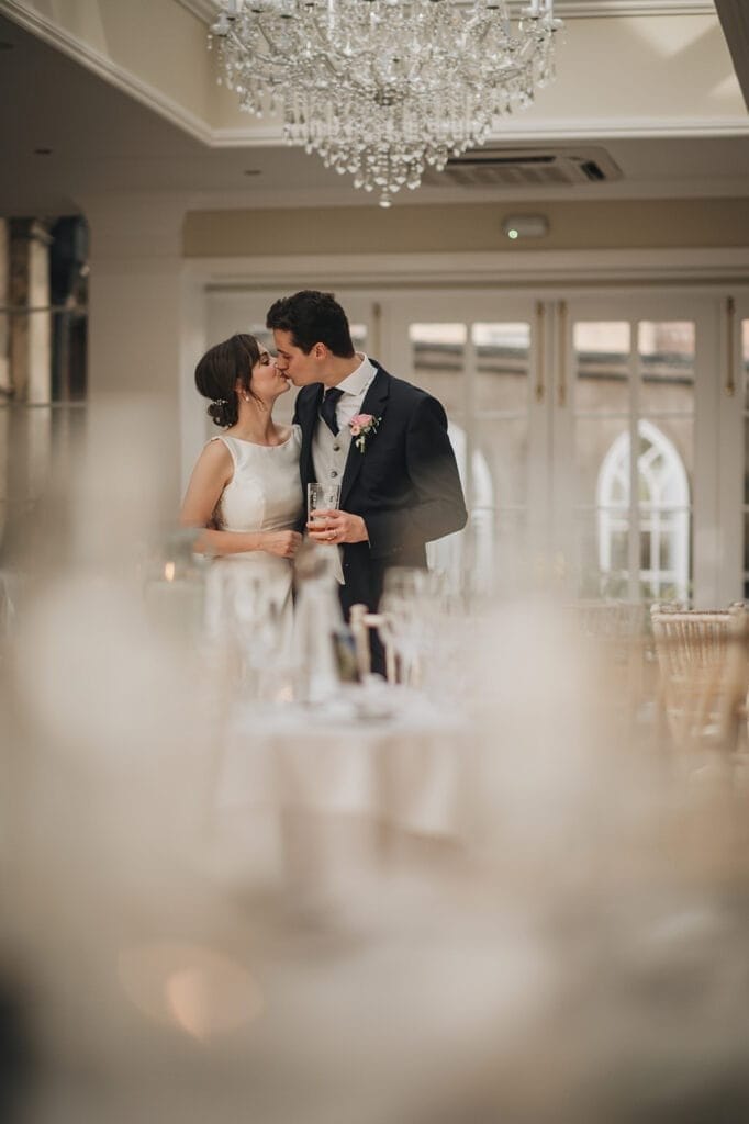 Goldsborough Hall Wedding | Yorkshire Wedding Photographer 61 A bride and groom stand close, smiling under a chandelier in an elegant room at Goldsborough Hall. The groom holds a drink, and the couple appears joyful. The foreground is softly blurred, highlighting their intimate moment captured by the wedding photographer.