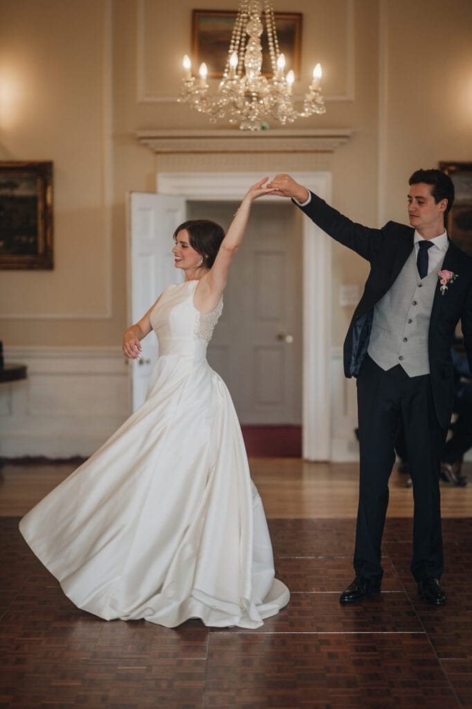 Goldsborough Hall Wedding | Yorkshire Wedding Photographer 77 A bride in a flowing white wedding dress and a groom in a dark suit dance gracefully at Goldsborough Hall, their elegance mirrored by chandeliers and framed paintings. As she twirls, the moment is perfectly captured by a skilled wedding photographer.