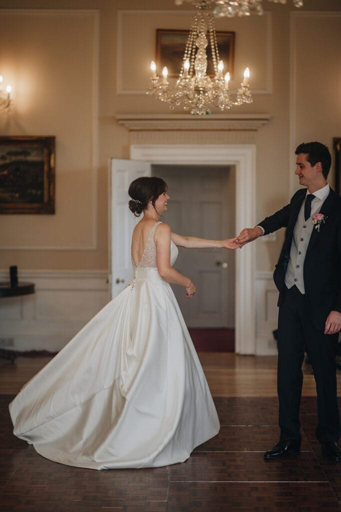 Goldsborough Hall Wedding | Yorkshire Wedding Photographer 78 A bride in a flowing white wedding dress and a groom in a dark suit dance gracefully at Goldsborough Hall, their elegance mirrored by chandeliers and framed paintings. As she twirls, the moment is perfectly captured by a skilled wedding photographer.