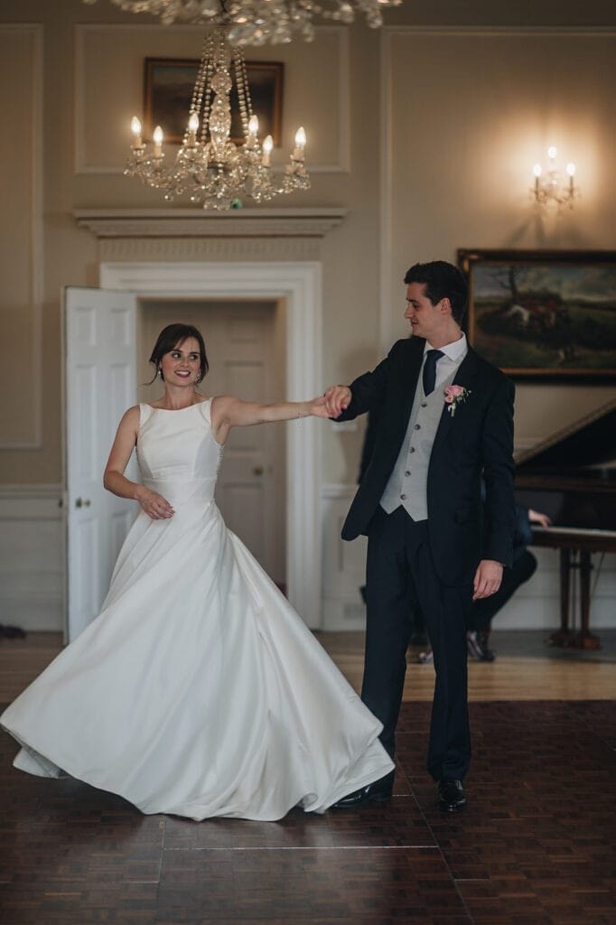 Goldsborough Hall Wedding | Yorkshire Wedding Photographer 79 A couple dances gracefully in a formal setting at Goldsborough Hall. The woman wears a flowing white gown, while the man is in a dark suit with a vest and tie. A chandelier hangs above them, captured exquisitely by their wedding photographer, as a piano is visible in the background.
