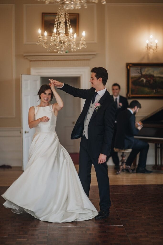 Goldsborough Hall Wedding | Yorkshire Wedding Photographer 80 A couple dances gracefully in a formal setting at Goldsborough Hall. The woman wears a flowing white gown, while the man is in a dark suit with a vest and tie. A chandelier hangs above them, captured exquisitely by their wedding photography, as a piano is visible in the background.