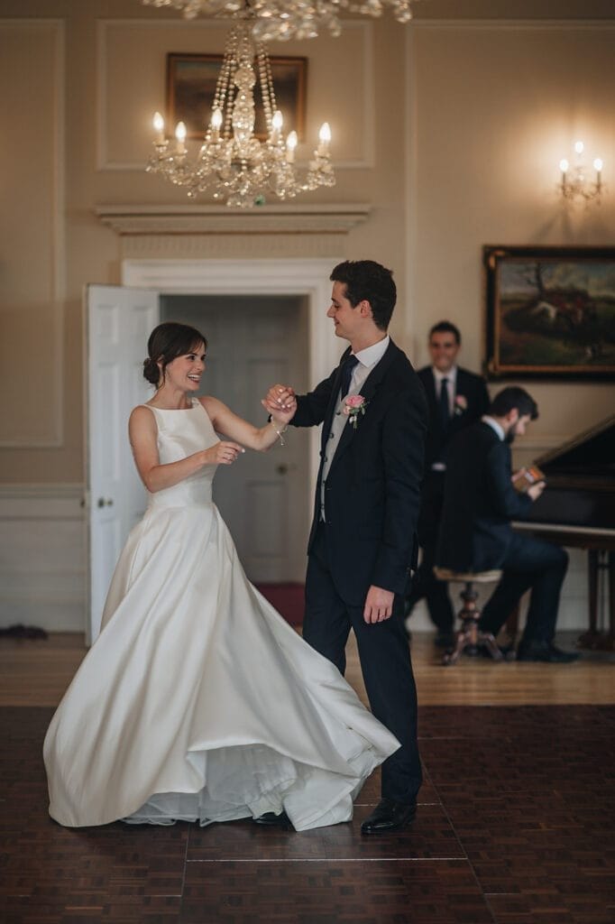 Goldsborough Hall Wedding | Yorkshire Wedding Photographer 81 A bride in a white gown and a groom in a black suit joyfully dance beneath the chandelier at Goldsborough Hall. A man in a suit plays the piano softly, capturing the elegance and warmth of this enchanting moment through their wedding photographers lens.
