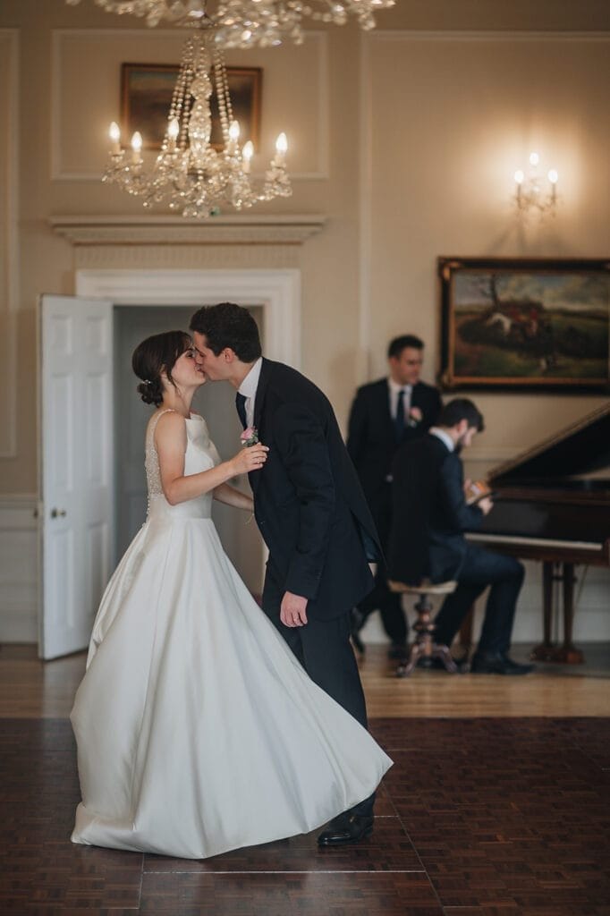 Goldsborough Hall Wedding | Yorkshire Wedding Photographer 82 A couple dressed in wedding attire shares a kiss while dancing in an elegant room at Goldsborough Hall. A pianist and a standing man linger in the background as chandeliers illuminate their joy, with a stunning painting adorning the wall—a scene worthy of any wedding photographers lens.