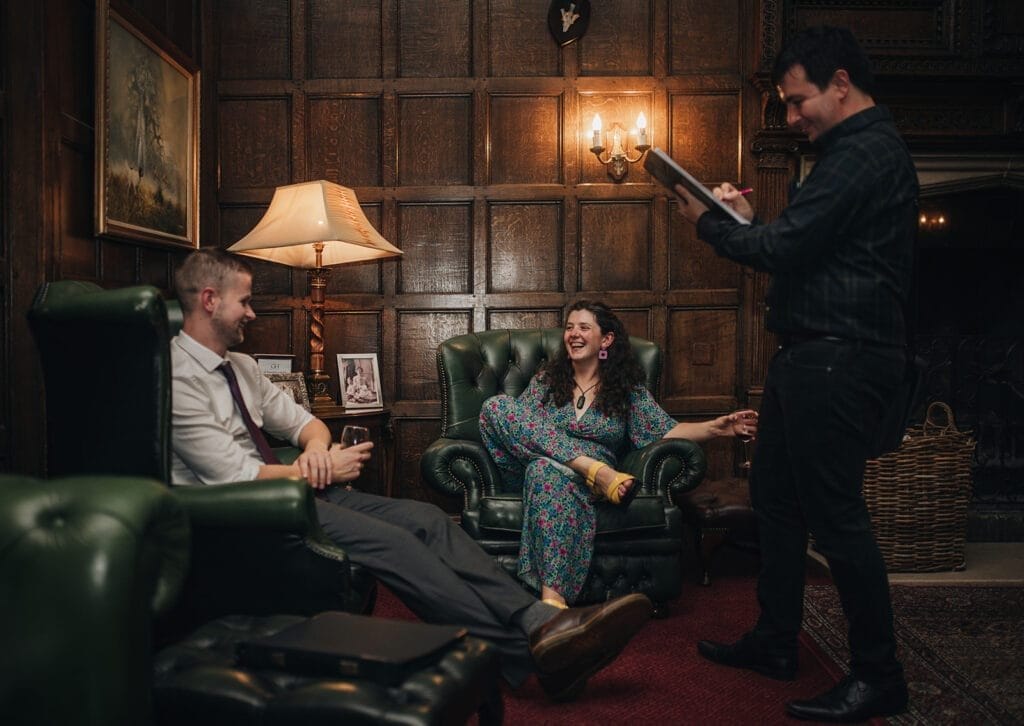Goldsborough Hall Wedding | Yorkshire Wedding Photographer 113 In a cozy, wood-paneled room, reminiscent of Goldsborough Hall, three people converse. One sits in a green armchair chatting while another sits cross-legged in another armchair, smiling widely. A third stands jotting notes on a notepad. A lamp and framed photo adorn the side table, adding warmth to the scene.