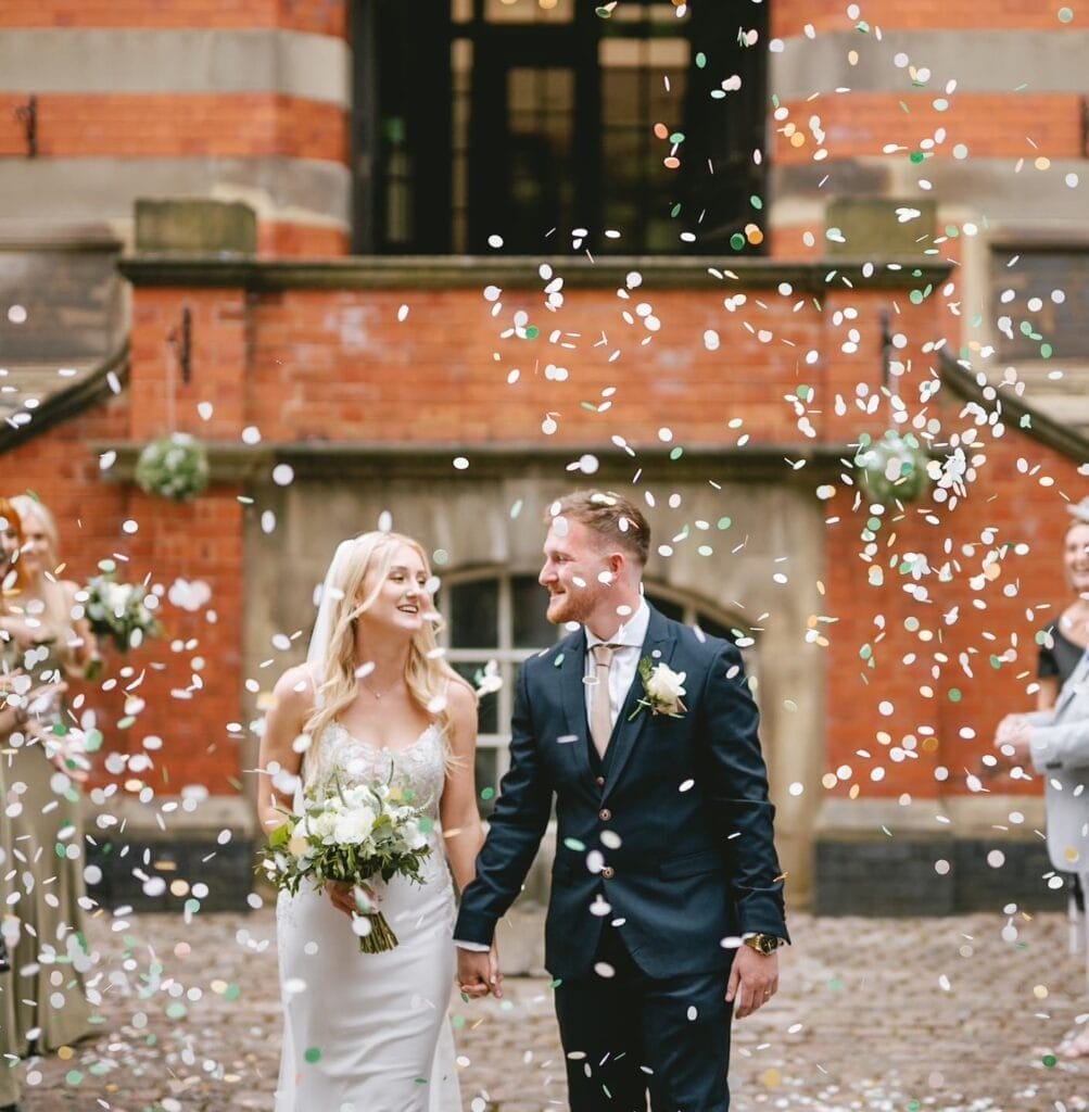 The bride and groom smile at each other, hand in hand, amidst falling white confetti. She holds a bouquet as their loved ones celebrate outside the charming red brick backdrop of the Pumping House Wedding Venue.