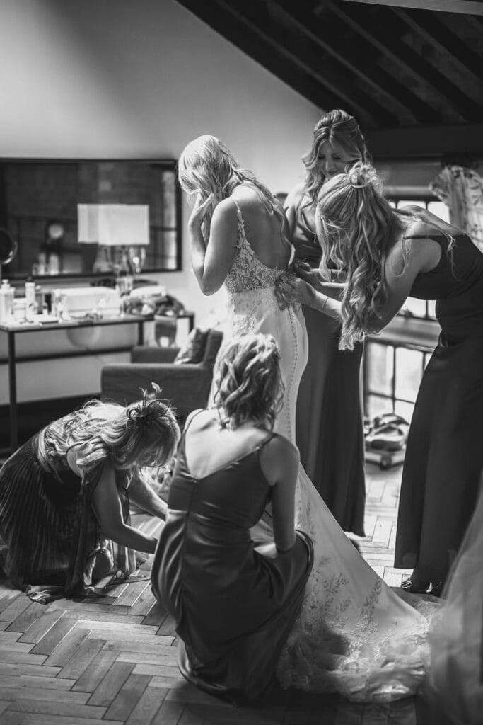 In a serene room at the Pumping House wedding venue, a bride stands with her back to the camera in a lacy white gown. Four bridesmaids assist her with the dress and veil. The rustic wooden floor and dressing table create an elegant atmosphere in this timeless black and white photo.