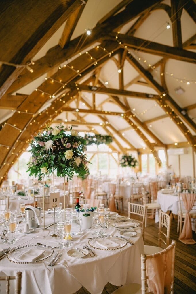 The beautifully decorated wedding reception hall, perfect for an LGBTQ+ celebration, features wooden beams and elegantly set tables. Chairs adorned with white linens complement tall floral centerpieces, while warm ambient string lights add charm to the festive atmosphere.
