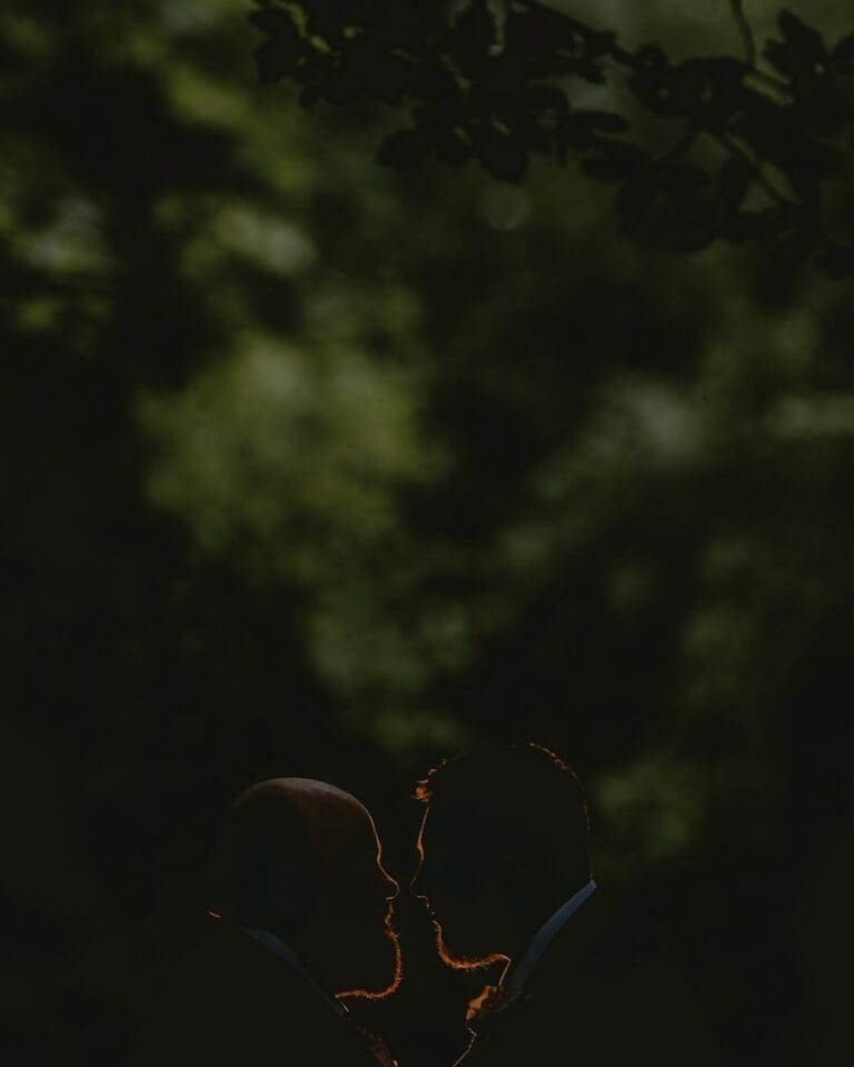 Silhouette of two people facing each other against a dark, leafy backdrop, with subtle lighting highlighting their profiles—a moment beautifully captured by Sandburn Hall wedding photographer.