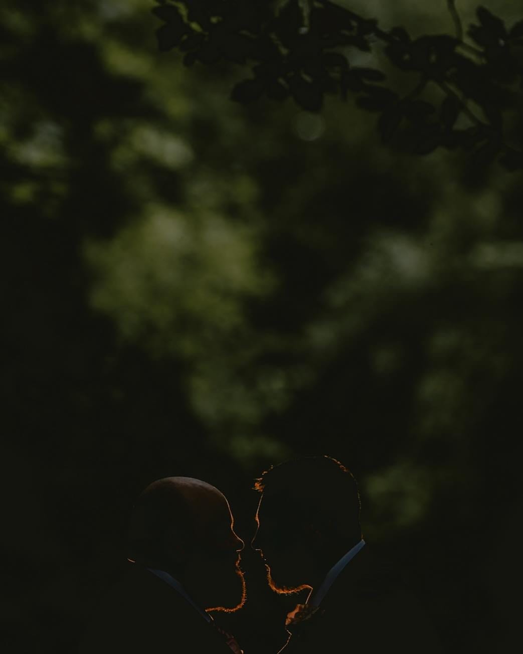 Silhouette of two people facing each other against a dark, leafy backdrop, with subtle lighting highlighting their profiles—a moment beautifully captured by Sandburn Hall wedding photographer.