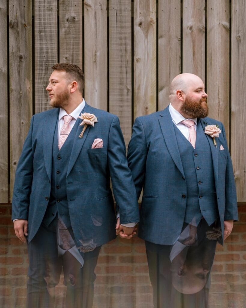 Two men in blue suits with pink ties and floral boutonnieres stand back-to-back, holding hands. In front of a wooden fence, they gaze in opposite directions, perfectly capturing the essence of an LGBTQ+ wedding.