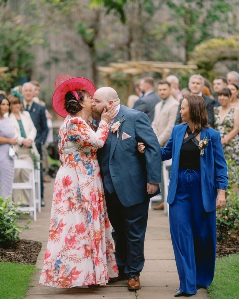 At an LGBTQ+ wedding, a bearded groom in a blue suit is kissed on the cheek by a woman in a floral dress and red hat, while another woman in a blue outfit smiles. Guests sit and stand in the background during an outdoor ceremony captured by Sandburn Hall wedding photographer.