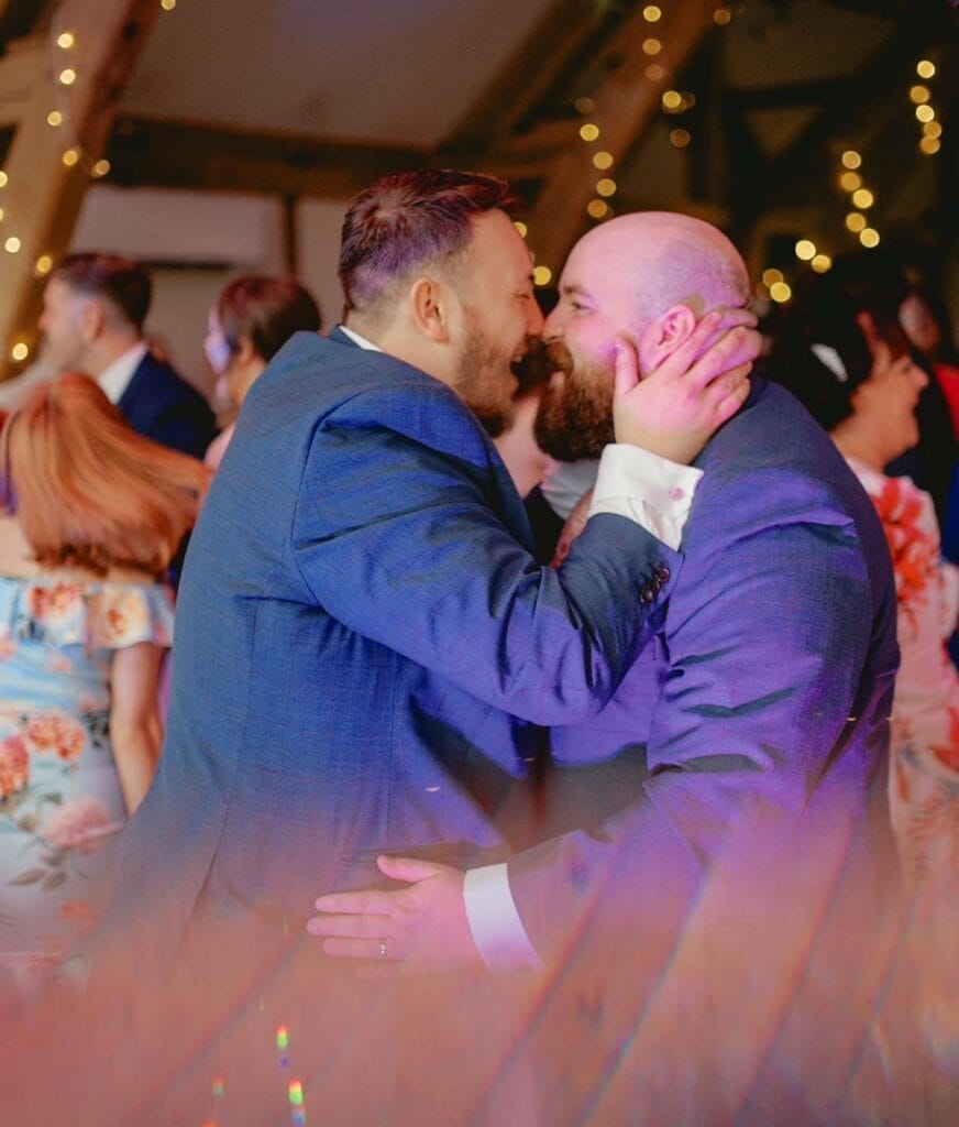 Two men in suits share an affectionate moment at an indoor event, resembling a beautiful LGBTQ+ wedding. They embrace happily, surrounded by softly lit strings of lights and guests in formal attire, reminiscent of scenes captured by a Sandburn Hall wedding photographer.