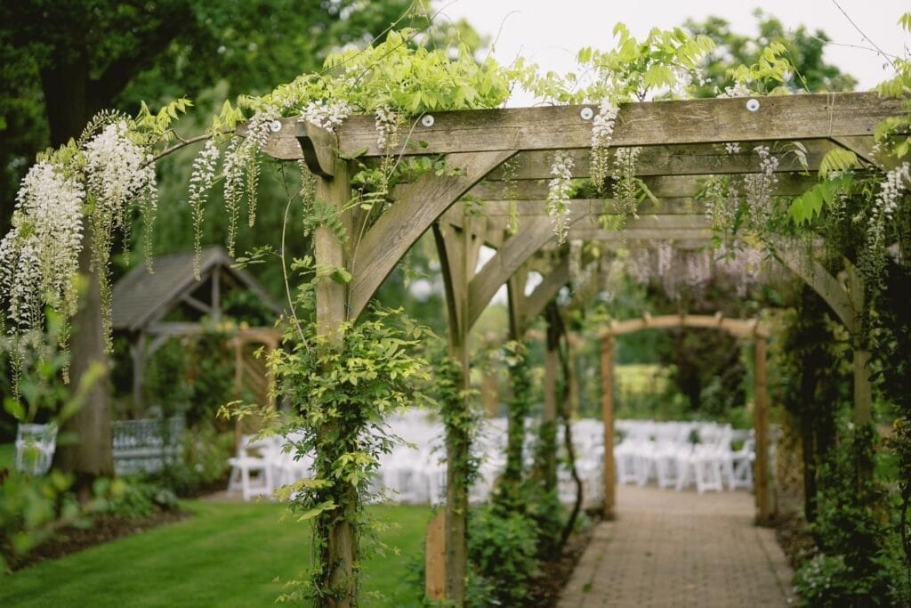 A wooden pergola adorned with lush green vines and white flowers extends over a brick pathway. White chairs are arranged in the background, suggesting an LGBTQ+ wedding setting, surrounded by greenery and trees. Perfectly capturing these moments is a Sandburn Hall wedding photographers dream.