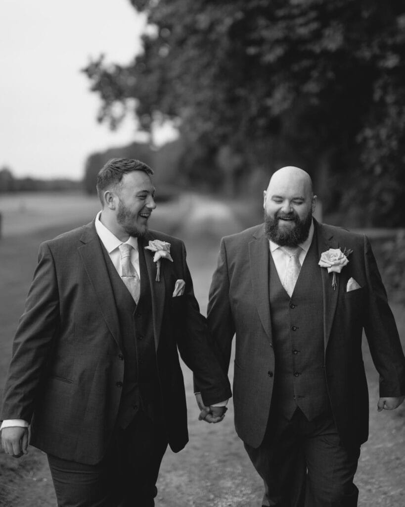 Two men in suits walk hand in hand along a tree-lined path, celebrating their LGBTQ+ wedding. Each wears a boutonnière with light-colored flowers, smiling joyfully against the blurred natural backdrop—a picturesque moment perfectly captured by Sandburn Halls skilled wedding photographer.